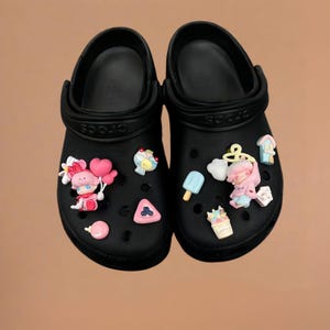 May include: A pair of black Crocs sandals decorated with colorful charms. The charms include a pink character holding balloons, ice cream, a cloud, and other cute designs. The sandals are shown on a neutral background.