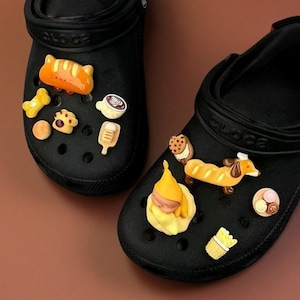 May include: Black clogs adorned with colorful food-themed charms. Charms include a baguette, coffee cup, cookie, bone, and a sleeping gnome. The clogs are decorated with various food-related accessories.