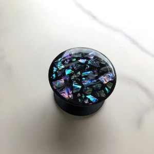May include: A black ear gauge with a domed, iridescent abalone shell inlay. The shell pieces reflect shades of blue, green, and purple. The gauge is round and has a flared base.