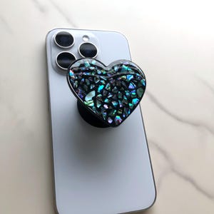May include: A heart-shaped phone grip with a black base and a mosaic design of iridescent, dark blue, and green pieces. The grip is attached to the back of a white smartphone.