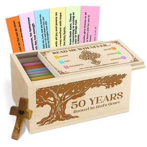May include: A wooden box with a tree design and the text "50 YEARS Rooted in God's Grace." The box contains colorful cards with Bible verses. A wooden cross with the name "Lucas" is also present.