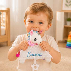 May include: A white plush unicorn security blanket featuring a textured fabric body with the name Emma embroidered in blue script. The toy includes a colorful striped mane, a patterned horn, a pink heart accent, and a white star-shaped plastic teether.