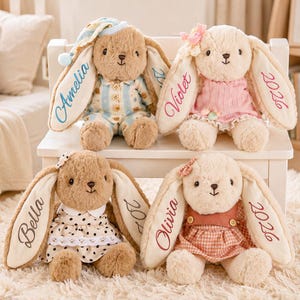 Peut inclure: Quatre lapins en peluche avec de longues oreilles portant des noms brodés et l'année 2026. Les lapins portent diverses tenues, notamment un pyjama à rayures bleues, une robe rose, une robe à pois blancs et une robe à carreaux rouille.