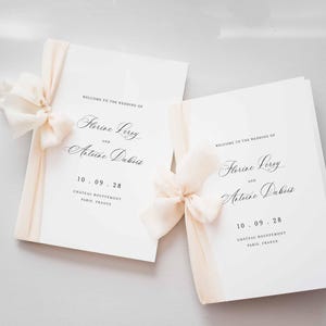 Pode incluir: Dois programas de casamento brancos com texto preto que diz Welcome to the wedding of Florine Leroy and Antoine Dubois, 10.09.28, Chateau Bouffemont, Paris, France. Cada programa é decorado com uma fita de chiffon pêssego pálido em laço.