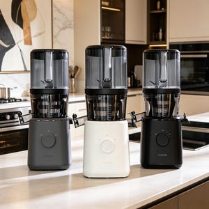 May include: Three contemporary juicers displayed in a kitchen. The juicers are in grey, white, and black. Each has a transparent top section and a base with a control dial. The brand name "nomo" is printed on the white and black juicers.