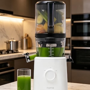 May include: A white Nama juicer with a clear container holding fruits and vegetables. A glass of green juice is beside the juicer. The juicer has a black top and a dial with "OFF" and "ON" settings. The word "nama" is printed on the front.