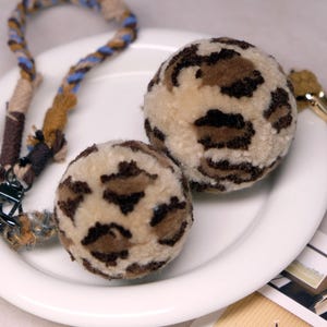 May include: Two plush dog toys with a leopard print pattern rest on a white plate. A matching rope dog leash with blue and brown accents is also present. The toys are round and fluffy, ideal for pet play.