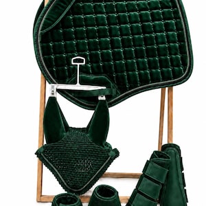 Op de afbeelding: Een donker smaragdgroene ruiterset bestaande uit een gewatteerd fluwelen zadeldek, een bijpassend gehaakt oornetje, twee hoefklokken en twee beenbeschermers. De items worden gepresenteerd op een houten rek en hebben een uniforme kleur.