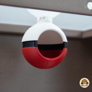 Puede incluir: Una carcasa esférica y hueca diseñada como una bola roja, blanca y negra con una gran abertura circular en la parte frontal. El objeto está unido a un soporte de ventosa blanco en la parte superior, lo que permite fijarlo a superficies lisas.