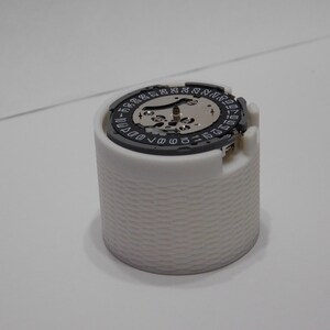 May include: A white, cylindrical watch component holder with a textured exterior. The top features a watch movement with a black and silver dial, displaying numbers and intricate gears. The overall design is clean and technical.