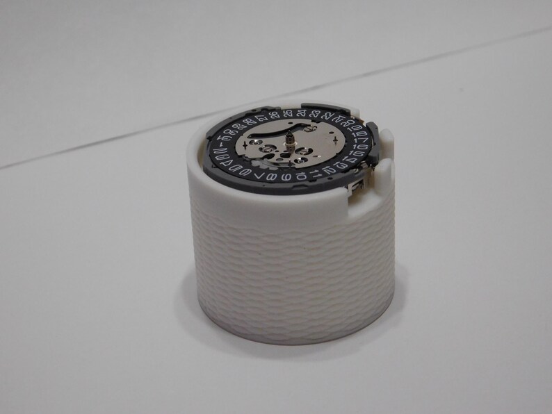 May include: A white, cylindrical watch component holder with a textured exterior. The top features a watch movement with a black and silver dial, displaying numbers and intricate gears. The overall design is clean and technical.