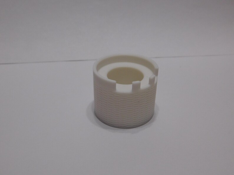 May include: A white, cylindrical object with a textured, basket-weave pattern on the exterior. The top has a circular opening and a crenelated edge, resembling a miniature castle. The object is likely a decorative or functional item.