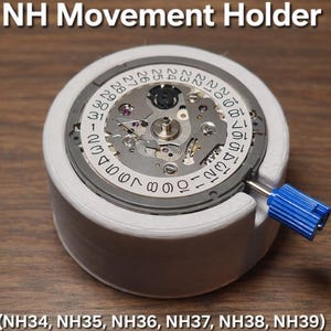 May include: A white NH Movement Holder with a watch movement inside. The watch movement has a silver face with black numbers and a blue tool. The text "NH Movement Holder" is at the top, and the model numbers are listed at the bottom.