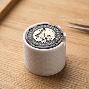 May include: A white cylindrical watch movement holder with a watch mechanism inside. The holder is on a wooden surface, with tweezers and a magnifying glass nearby. Text on the holder reads "PRECISION FDM PRINT - VK63 MOVT HOLDER".