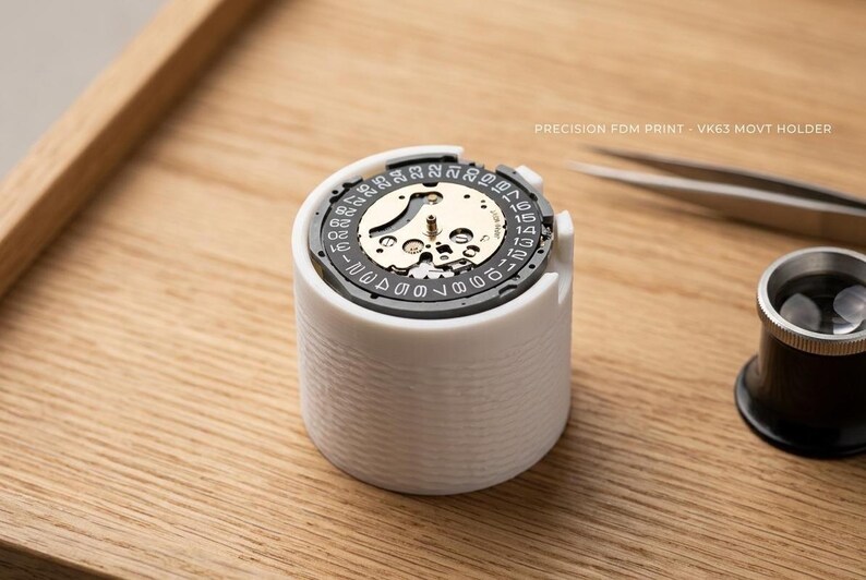 May include: A white cylindrical watch movement holder with a watch mechanism inside. The holder is on a wooden surface, with tweezers and a magnifying glass nearby. Text on the holder reads "PRECISION FDM PRINT - VK63 MOVT HOLDER".