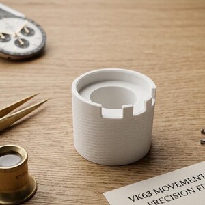 May include: A white, cylindrical watch movement holder with a castellated top, designed for precision FDM printing. The holder is on a wooden surface, alongside watch components and tools.