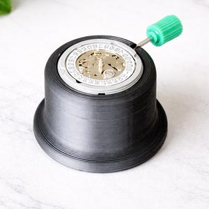 May include: A black watch repair tool with a watch movement inside. A green screwdriver is inserted into the watch. The tool has a wide base and a ridged exterior. The watch movement has gold and silver components.