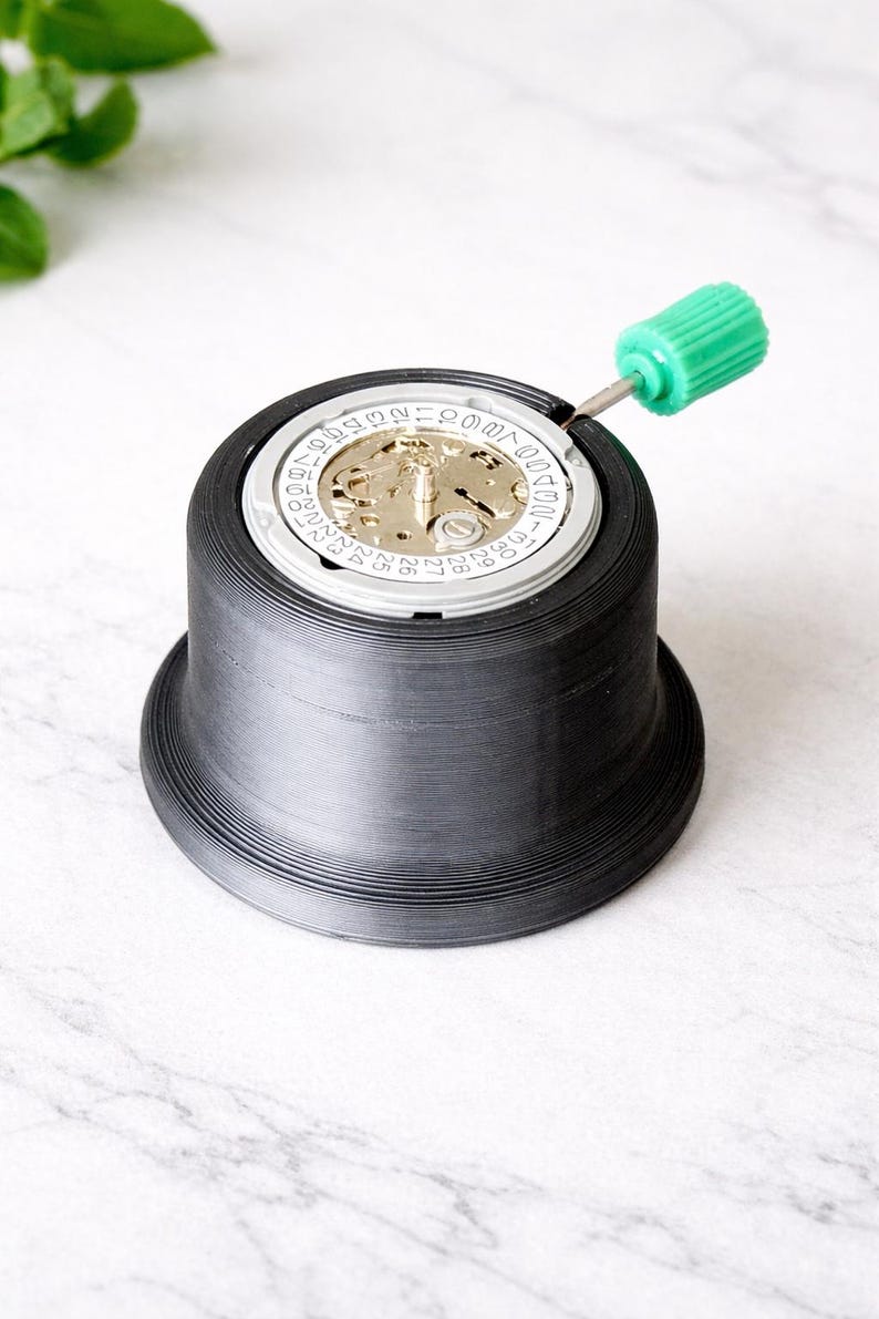 May include: A black watch repair tool with a watch movement inside. A green screwdriver is inserted into the watch. The tool has a wide base and a ridged exterior. The watch movement has gold and silver components.
