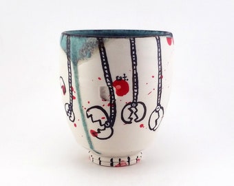 May include: A white ceramic cup with a teal rim and base. The cup is decorated with black line drawings of claw-like shapes hanging from red circles. Red paint splatters and drips add to the artistic design. The cup has a rounded shape.