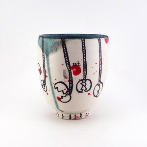 May include: A white ceramic cup with a teal rim and base. The cup is decorated with black line drawings of claw-like shapes hanging from red circles. Red paint splatters and drips add to the artistic design. The cup has a rounded shape.