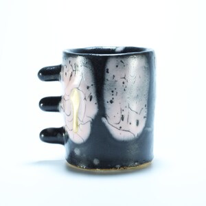 May include: Black ceramic mug with three finger-like handles and pink and white abstract designs.