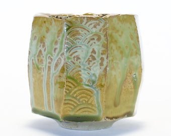 May include: A handcrafted ceramic bowl with a unique geometric shape. The bowl features a blend of green and gold glaze with a textured design. The design includes wave and tree patterns. The bowl is a decorative piece.