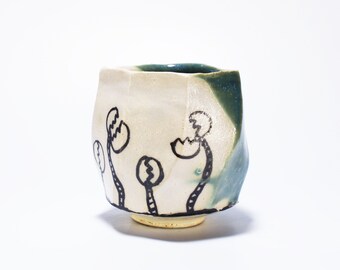 Puede incluir: Una taza pequeña de cerámica blanca con un esmalte verde en el lateral. La taza tiene una forma única e irregular y está decorada con diseños de flores estilizadas pintadas a mano en negro.