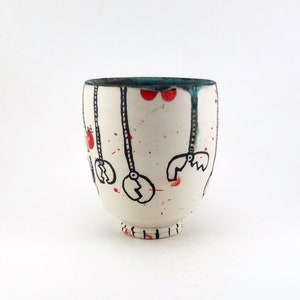 May include: A white ceramic cup with a unique design. The cup features black line art of hanging objects, red splatters, and red circles. The rim is teal, and the base has a small foot. The cup is a handmade piece.