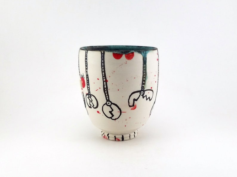 May include: A white ceramic cup with a unique design. The cup features black line art of hanging objects, red splatters, and red circles. The rim is teal, and the base has a small foot. The cup is a handmade piece.