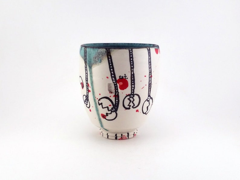 May include: A white ceramic cup with a teal rim and base. The cup is decorated with black line drawings of claw-like shapes hanging from red circles. Red paint splatters and drips add to the artistic design. The cup has a rounded shape.