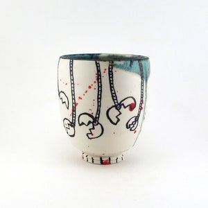 May include: A white ceramic cup with a rounded body and a small foot. The cup is decorated with black line drawings of claw-like shapes hanging from dotted lines, with red accents. The rim has a blue-green glaze.