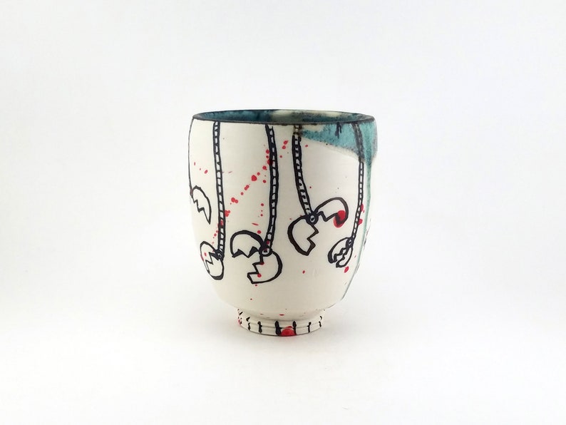 May include: A white ceramic cup with a rounded body and a small foot. The cup is decorated with black line drawings of claw-like shapes hanging from dotted lines, with red accents. The rim has a blue-green glaze.