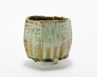 May include: A small, green and brown ceramic cup with a textured, irregular surface. The cup has a slightly rounded shape and a wide, open mouth.
