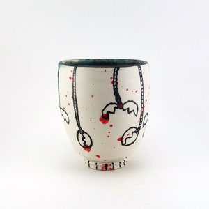 May include: A white ceramic cup with a dark teal interior and a black and red design. The cup features black line drawings with red splatter accents. The base of the cup has a series of small, horizontal lines.