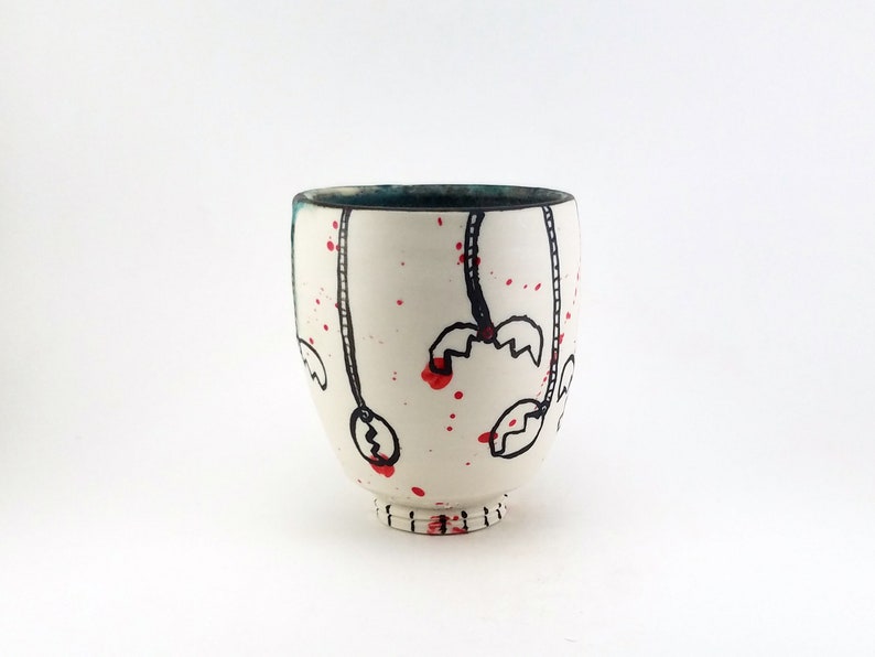 May include: A white ceramic cup with a dark teal interior and a black and red design. The cup features black line drawings with red splatter accents. The base of the cup has a series of small, horizontal lines.