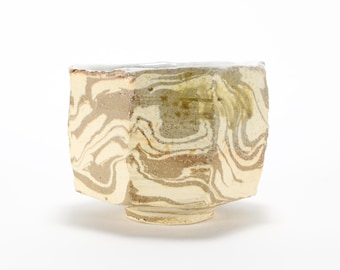 Puede incluir: Una taza de té de cerámica blanca con un diseño marmoleado en tonos marrones y beige. La taza tiene una forma ligeramente redondeada y una base ancha y plana.