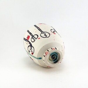 May include: A white ceramic cup with black, red, and blue abstract designs. The cup features black line art with red accents and red paint splatters. The bottom of the cup has a blue and green spiral design.