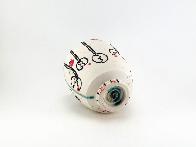 May include: A white ceramic cup with black, red, and blue abstract designs. The cup features black line art with red accents and red paint splatters. The bottom of the cup has a blue and green spiral design.
