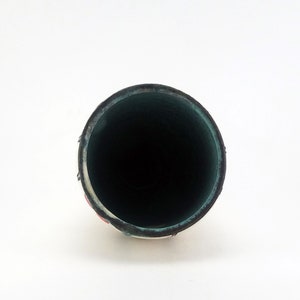 May include: A cylindrical, ceramic vase with a dark interior and a white exterior with red and black accents. The rim of the vase is dark green. The vase is set against a white background.