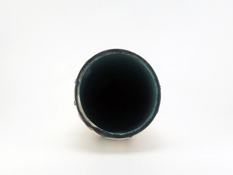 May include: A cylindrical, ceramic vase with a dark interior and a white exterior with red and black accents. The rim of the vase is dark green. The vase is set against a white background.