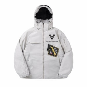 May include: A light grey hooded jacket with a black lining. The jacket features the text "Vera Vachetta" and a "GORE-TEX" patch. It has a zip closure and multiple pockets. The jacket is designed for outdoor activities.