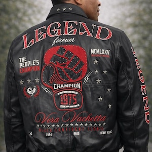 Może przedstawiać: Czarna skórzana kurtka z czerwono-białymi haftowanymi detalami. Tył przedstawia napis "LEGEND" i grafikę rękawicy bokserskiej. Dodatkowy tekst zawiera "The People's Champion" i "1975".