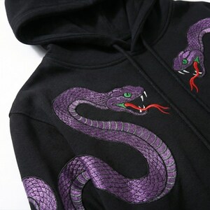 May include: Black hooded sweatshirt with embroidered purple snakes. The snakes have green eyes and red tongues, creating a striking contrast. The design is repeated on the front and sleeve. The hood is visible.