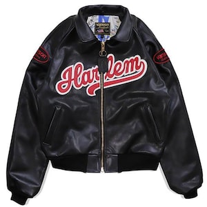 Peut inclure: Veste en cuir noir avec le mot "Harlem" en rouge sur la poitrine. La veste a une fermeture éclair dorée, deux poches avant et des écussons rouges sur les manches avec le mot "Vanson". La doublure intérieure a un motif.