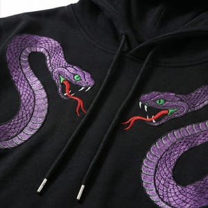 May include: Black hooded sweatshirt with two embroidered purple snakes, green eyes, and red tongues. The snakes are on the front, creating a striking design. The hoodie has a drawstring. The design is detailed.