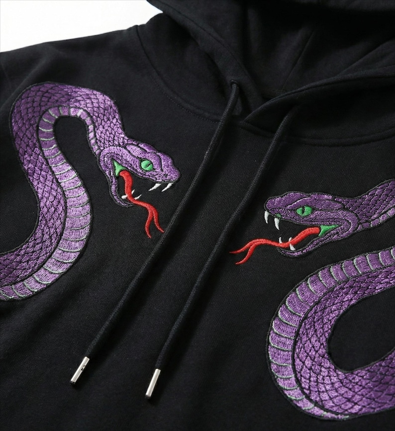 May include: Black hooded sweatshirt with two embroidered purple snakes, green eyes, and red tongues. The snakes are on the front, creating a striking design. The hoodie has a drawstring. The design is detailed.