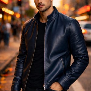 May include: A navy blue leather jacket with a zip-up front and a stand-up collar. The jacket is worn over a black t-shirt and paired with dark jeans. The model is standing on a city street.