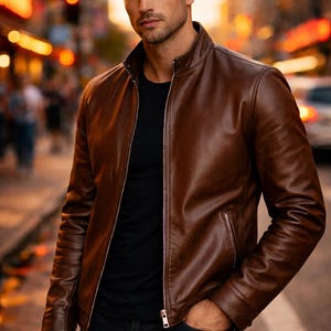 May include: A brown leather jacket with a zip-up front and a stand-up collar. The jacket is worn over a black t-shirt and paired with black jeans. The background shows a city street with blurred lights.