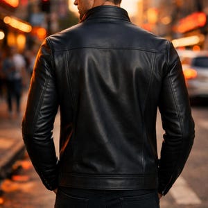 May include: A black leather jacket with a stand-up collar and a tailored fit. The jacket has a classic design with visible stitching and a smooth, glossy finish. The person is wearing black jeans. The background is a blurred city street.