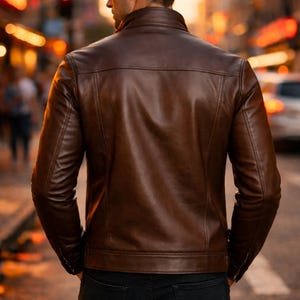 May include: A brown leather jacket with a classic design. The jacket features a stand-up collar, a front zipper, and pockets. The person is wearing black jeans. The background is a blurred city street.